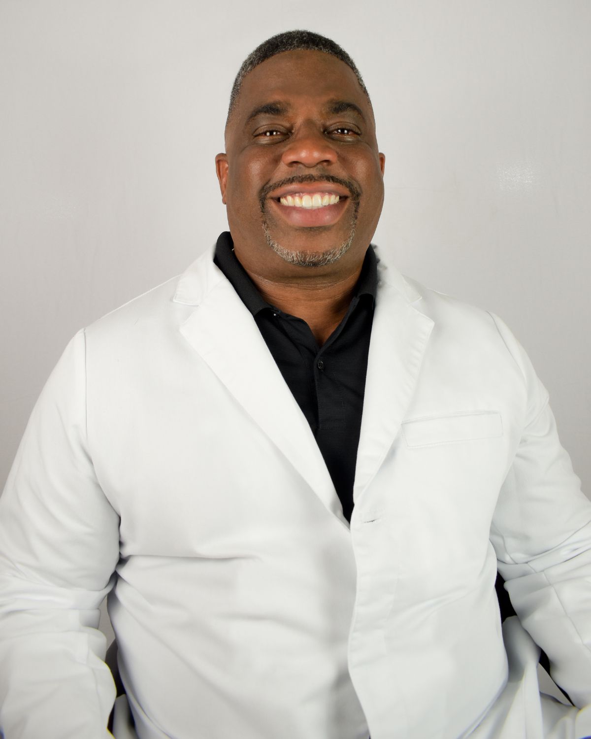 Professional portrait of smiling dentist wearing white coat against neutral background.