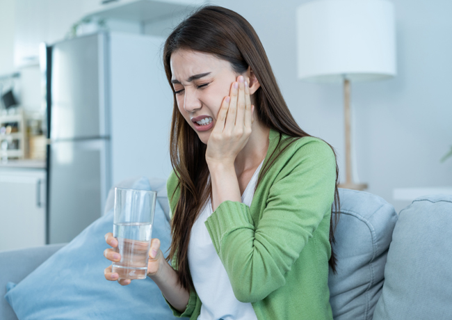 Woman holding water glass touching her cheek, appearing to experience tooth pain in home setting.