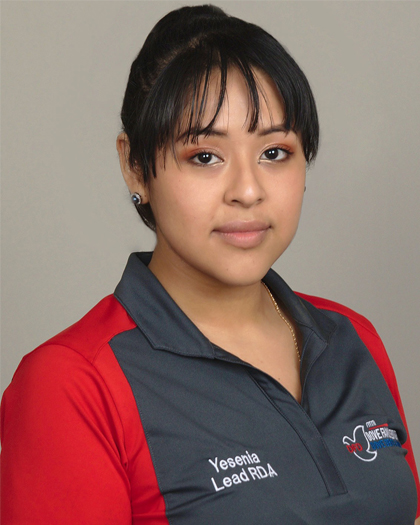 Young female dental staff member in red and navy scrubs against neutral background.