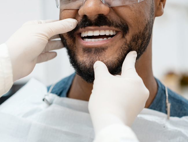 Man smiling during dental examination with gloved dentist hands checking teeth in clinical setting.