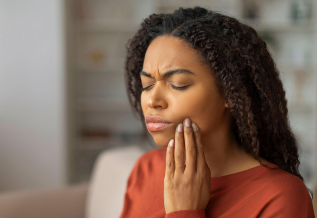Woman in red shirt touching her cheek, appearing to experience tooth or jaw pain indoors.