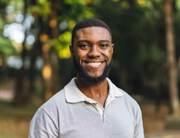 Smiling man in light gray polo shirt outdoors with blurred green trees in background.
