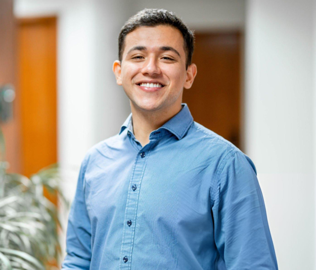 Professional headshot of smiling male dentist wearing blue shirt in modern dental office hallway.