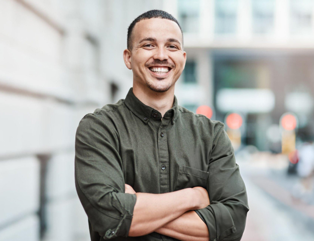Smiling man with crossed arms wearing olive button-up shirt in urban outdoor setting.