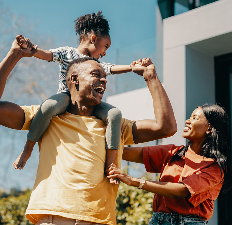 Happy family playing outdoors with father holding child on shoulders, mother smiling beside them.