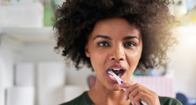 Woman brushing teeth in bathroom, smiling at camera with toothbrush in mouth.
