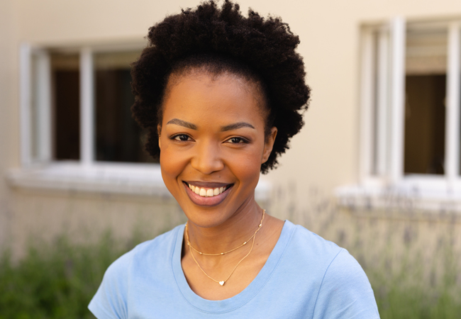 Smiling woman wearing light blue shirt outdoors in front of house with windows.