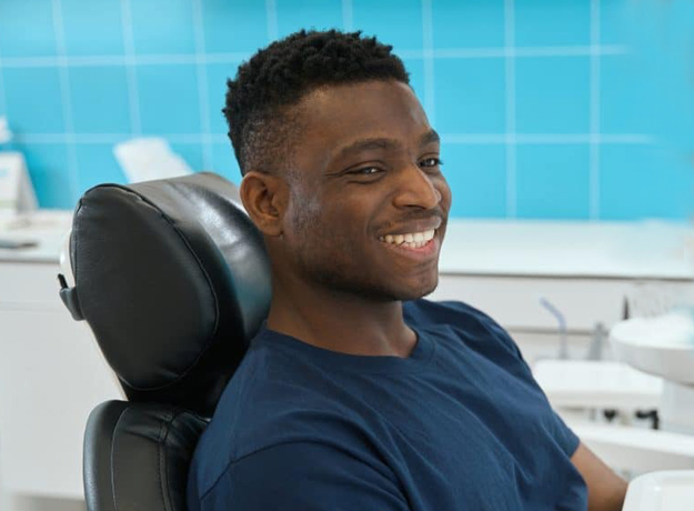 Smiling man sitting in dental chair at modern dentist office with blue background