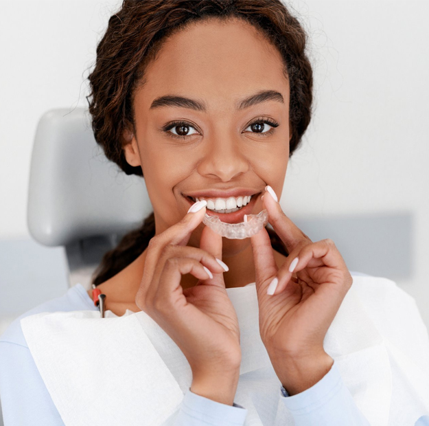 Woman holding clear dental aligner trays, smiling in clinical dental office setting.