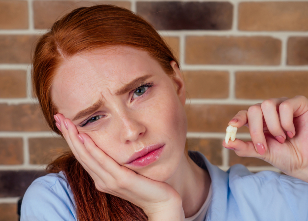 Young woman with red hair holding dental floss, looking frustrated against brick wall background.