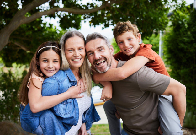 Happy family of four smiling outdoors in park setting with green trees.