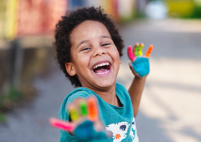 Happy young child with painted rainbow hands laughing outdoors in bright daylight.