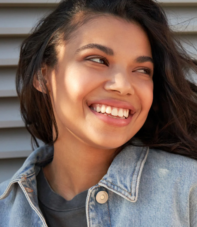 Young woman smiling brightly outdoors against wooden siding, wearing denim shirt.