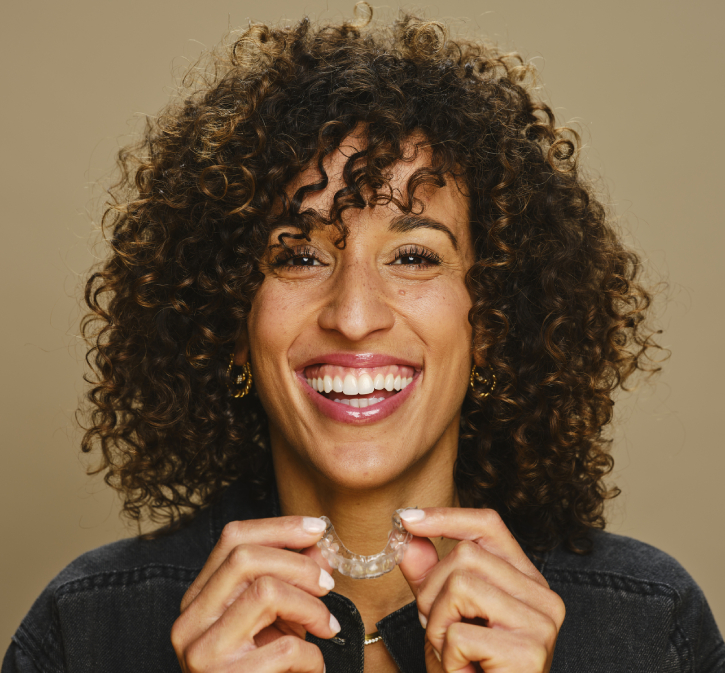 Woman with curly hair smiling while holding dental floss against neutral background.