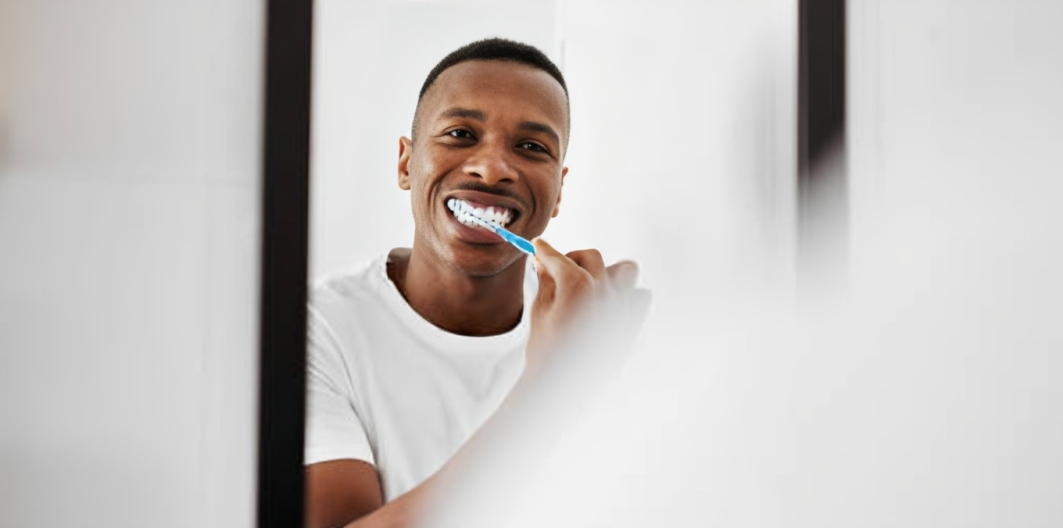 Man brushing teeth with white toothbrush in bathroom mirror, smiling.