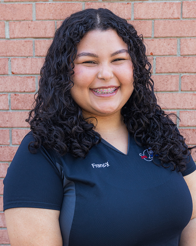 Professional headshot of smiling woman in navy Dove Family Dentistry polo against brick wall background.