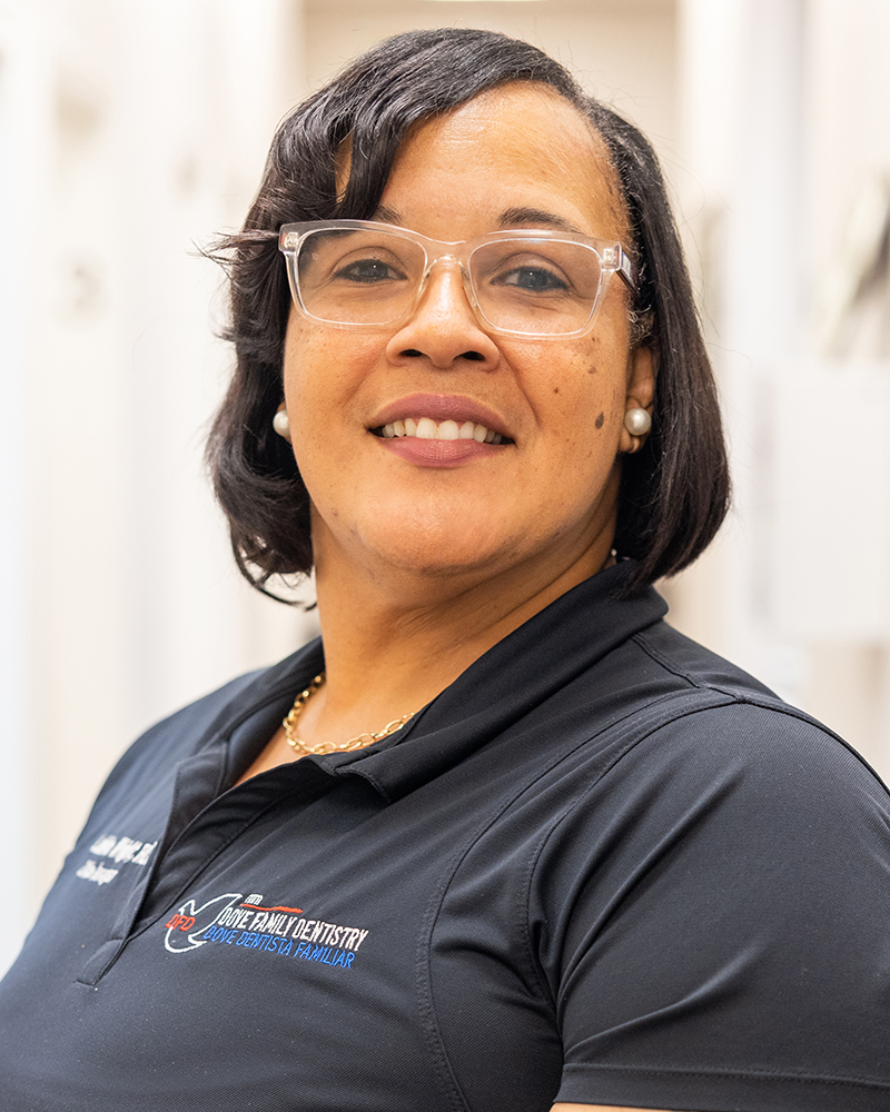 Woman wearing glasses and Dove Family Dentistry branded shirt, smiling at camera in professional office setting.