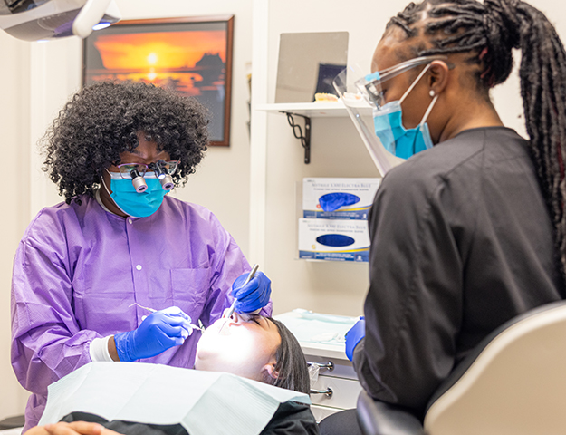 Dental hygienist in purple jacket examines patient's teeth in modern dental office treatment room.