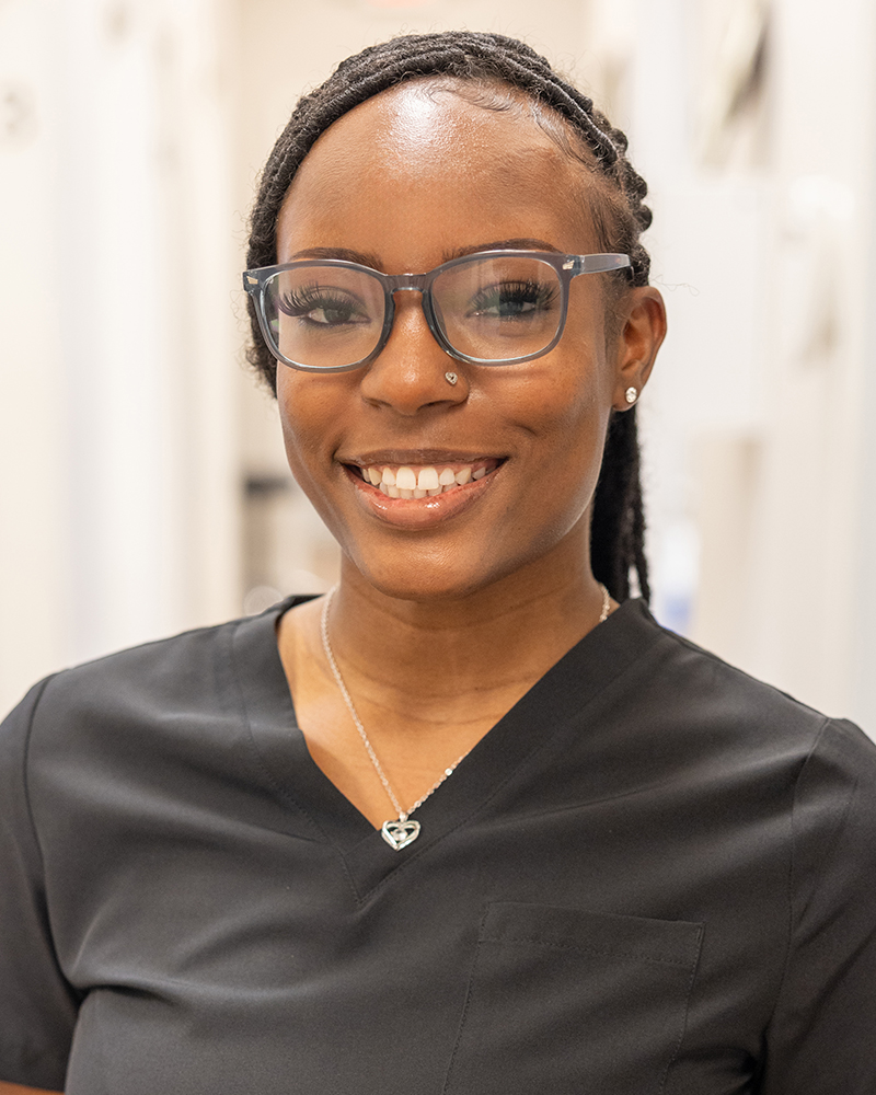 Female dental professional smiling at camera wearing glasses and black scrubs in clinical setting.