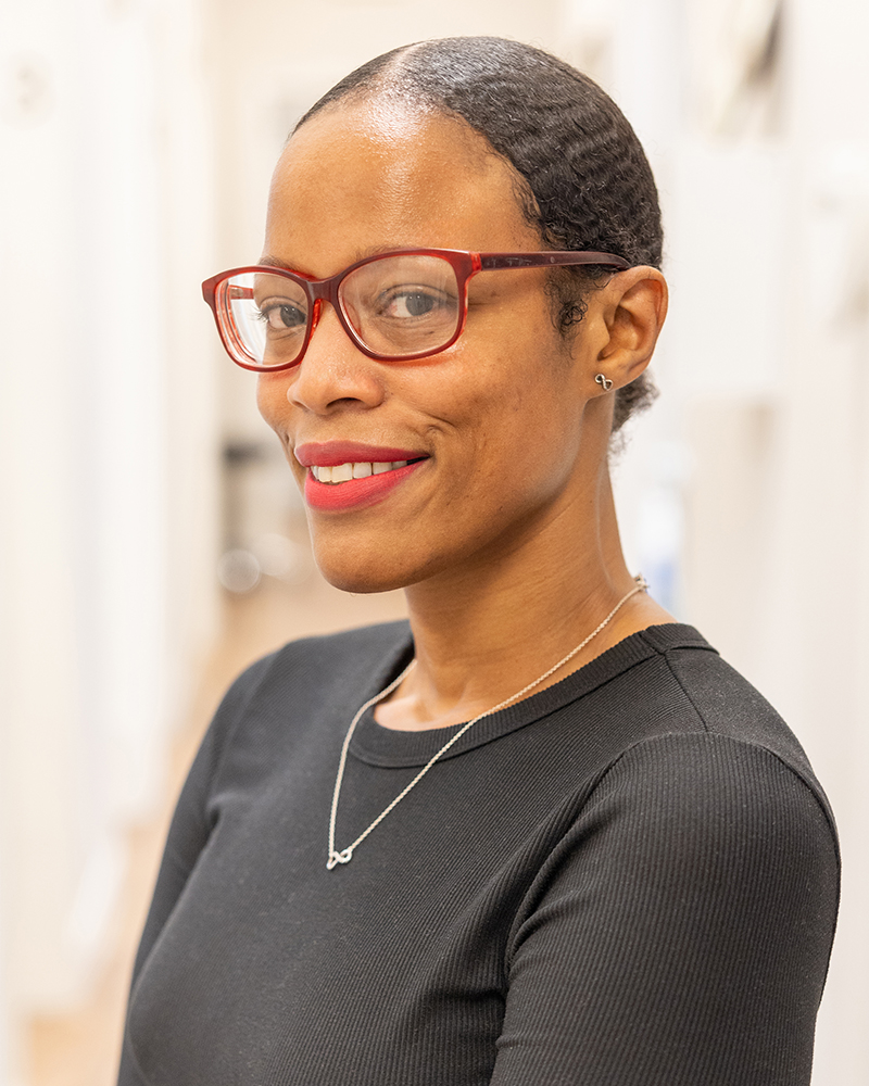 Professional woman wearing glasses and dark shirt smiles at camera in dental office setting.