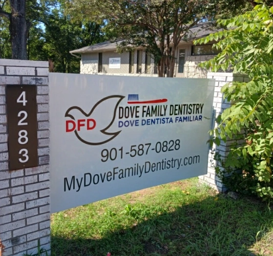 Dove Family Dentistry exterior sign with logo, phone number, and website on brick wall among trees.