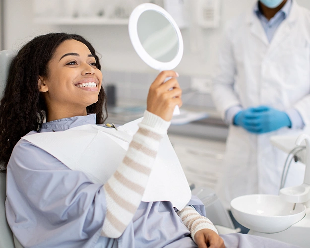 Woman admiring smile in dental mirror at dentistry clinic with dentist in background