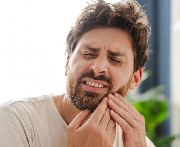 Man experiencing tooth pain, touching cheek with grimacing expression indoors.