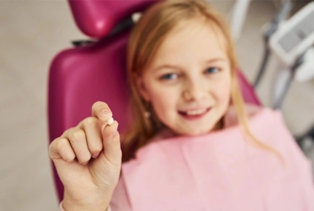 Young girl in pink shirt pointing at camera, sitting in dental chair at dentist's office.