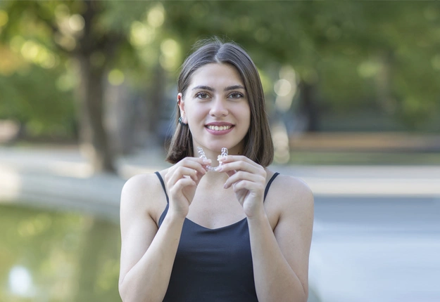 Woman smiling outdoors touching her teeth, park setting with blurred green background.