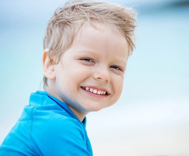 Happy young boy in blue shirt smiling at beach, dental health portrait.