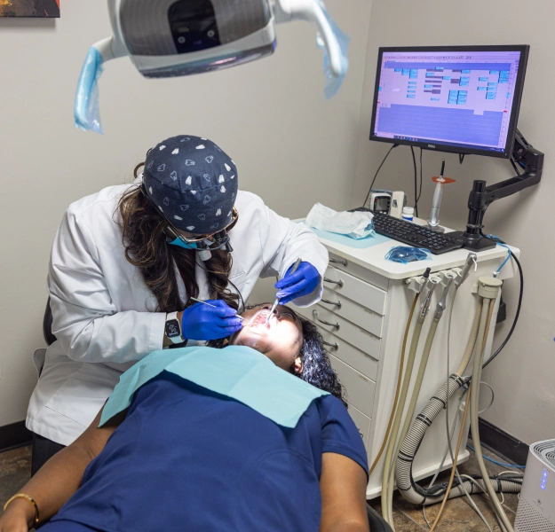 Dentist performing oral examination on patient in modern dental clinic operatory with overhead light and computer monitor.