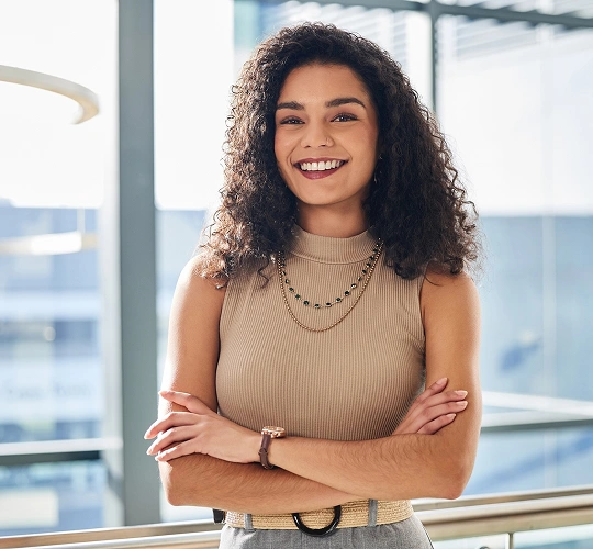 Professional woman smiling with crossed arms in modern office with city view windows.
