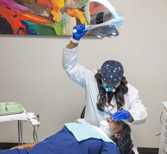 Dentist in protective gear examining patient's teeth in dental office treatment room.