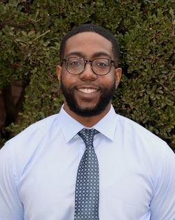 Professional headshot of smiling man in light blue shirt and tie against green foliage backdrop.