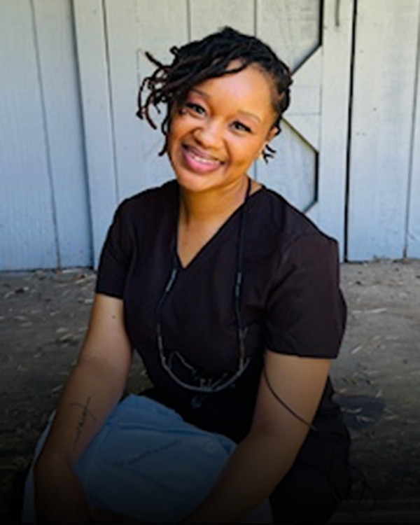 Smiling woman in black shirt posing against white corrugated metal wall backdrop.