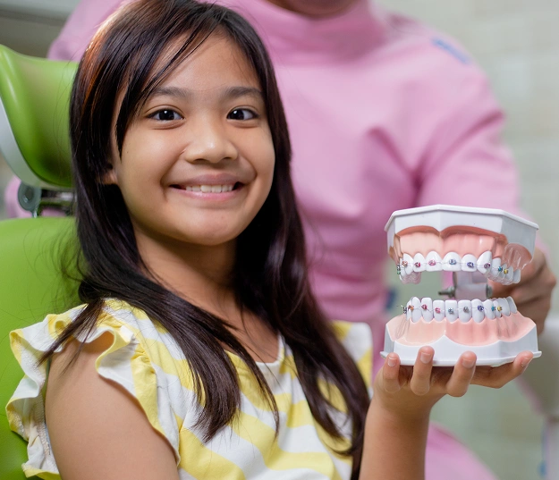 Young girl smiling while holding dental models showing braces in orthodontic office setting.