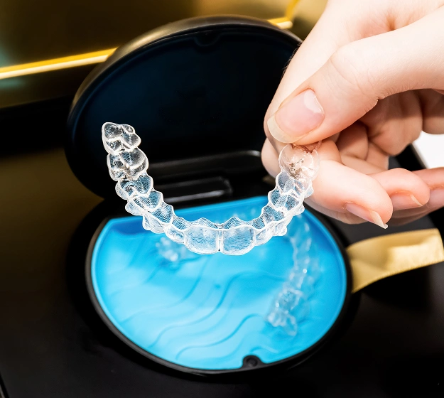 Person holding clear orthodontic aligner over blue dental mold tray in clinical setting.