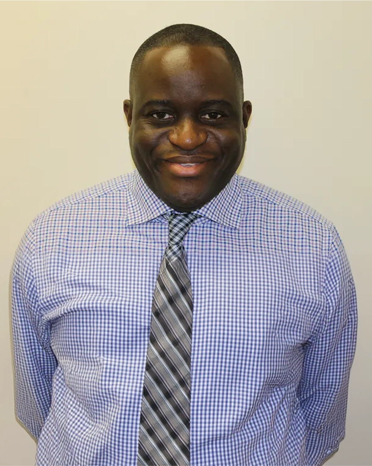 Professional headshot of smiling man in blue checkered shirt and striped tie against neutral background.