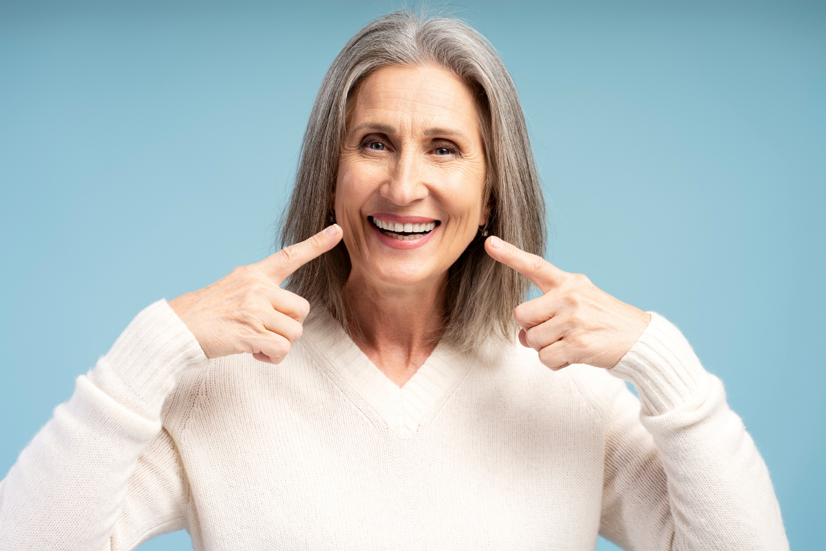 Woman with gray hair smiling and pointing to her teeth, demonstrating dental health results.